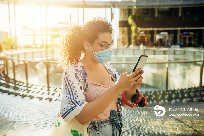 Young woman wearing surgical mask using smartphone outdoors