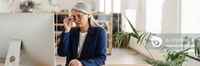 The Asian woman sitting at a table in front of the monitor adjusting glasses with her hand in the of