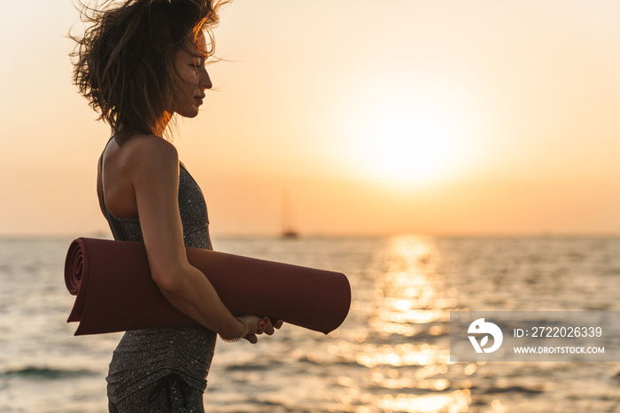 Photo of slim sportswoman looking away while standing with yoga mat by seaside in morning