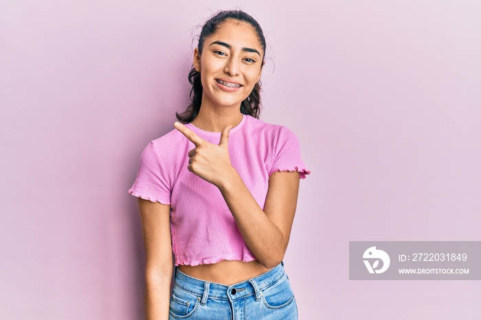 Hispanic teenager girl with dental braces wearing casual clothes smiling cheerful pointing with hand