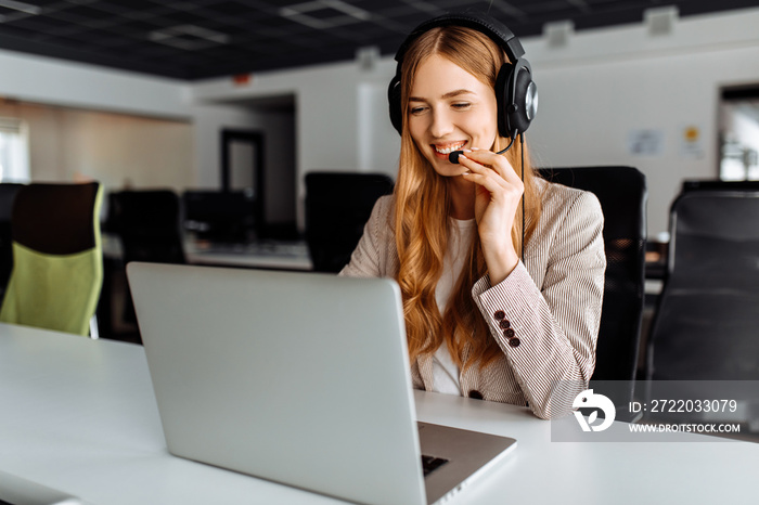 Smiling young female operator in headphones with headset working with laptop sitting at table at wor