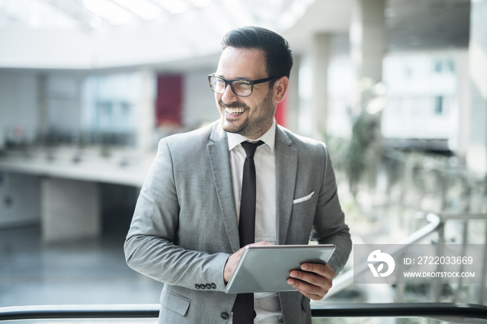 Smiling businessman looking away and using tablet while standing in business center hall.