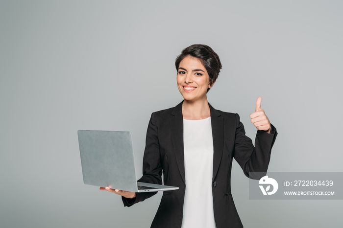 beautiful mixed race businesswoman holding laptop and showing thumb up isolated on grey