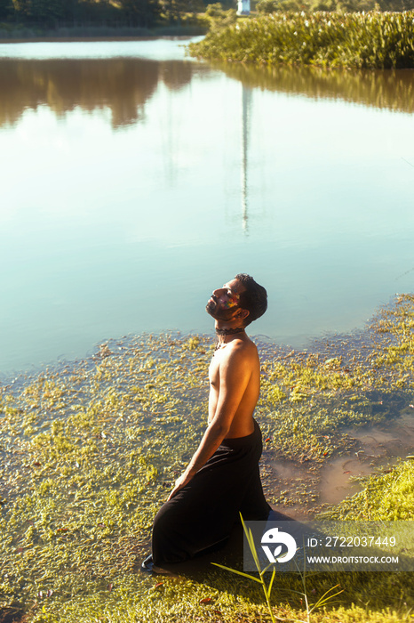 portrait of a dark skinned Indian man by a lake, shirtless, with make up, surrounded by nature