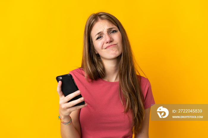 Young woman using mobile phone isolated on yellow background with sad expression