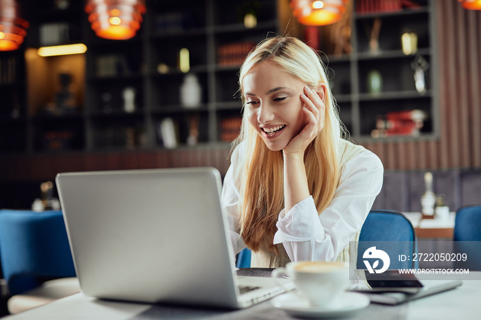 Beautiful smiling Caucasian blonde female blogger dressed smart casual sitting in cafeteria and usin