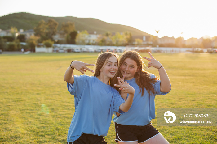 Portrait of happy girls showing peace sign while standing on soccer field against clear sky during s