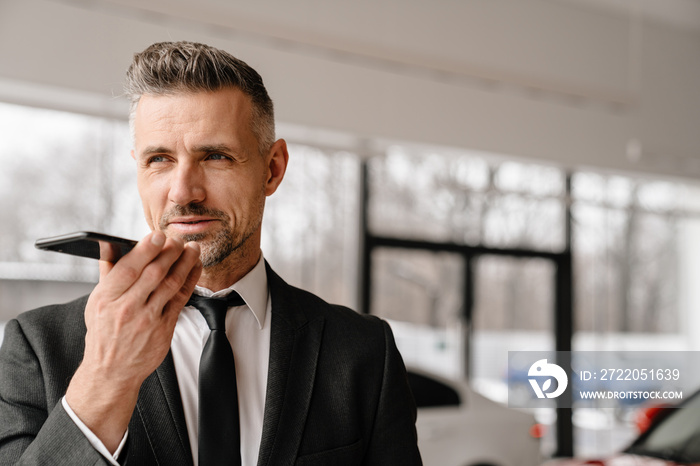 Mature white man talking on speakerphone while choosing car in showroom