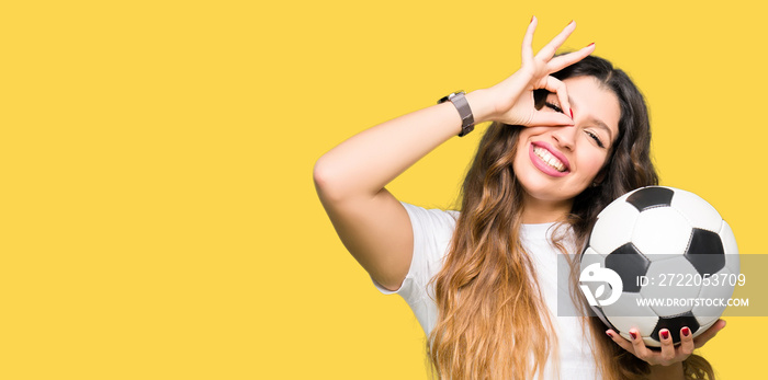 Young adult woman holding soccer football ball with happy face smiling doing ok sign with hand on ey