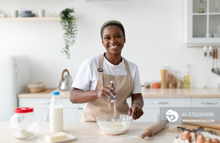 Smiling pretty millennial african american woman chef in apron making dough in light minimalist kitc