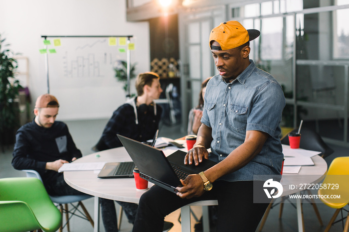 Dark skinned African american coworker in jeans shirt and yellow cap and using laptop in modern offi