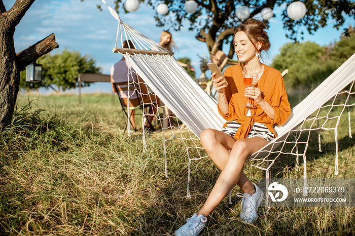 Young woman relaxing on the hammock, using smartphone during a lunch with friends in the garden on a