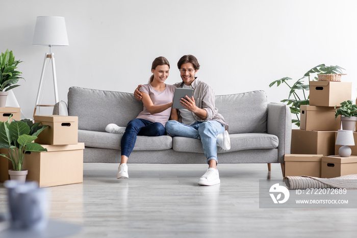 Smiling caucasian millennial guy show tablet to female sits on sofa in living room interior