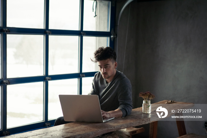 Handsome man using laptop computer on bench while sitting against window at home