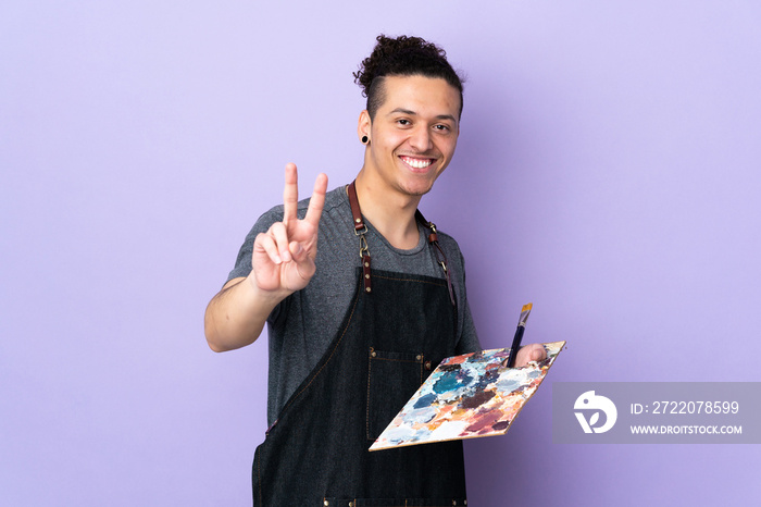 Young artist man holding a palette over isolated purple background smiling and showing victory sign