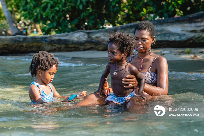 Horizontal de una madre junto a sus hijos afroamericanos sentados a la orilla de la playa bañándose 