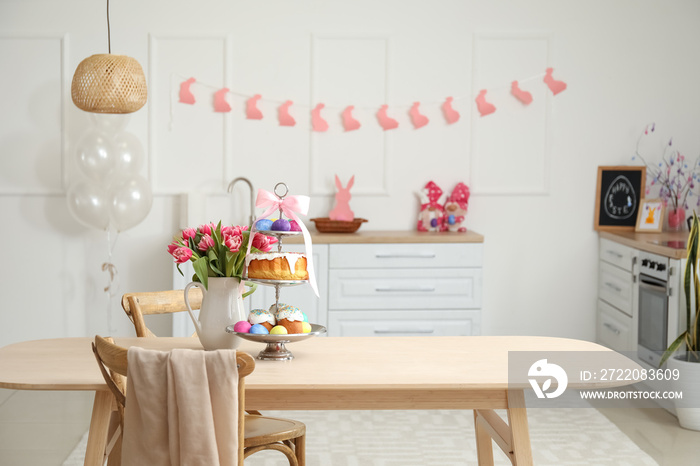 Stand with Easter cakes, eggs and tulips on dining table in kitchen