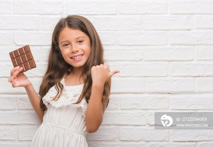 年轻的西班牙孩子在白色砖墙上吃着巧克力棒，用拇指指着并展示