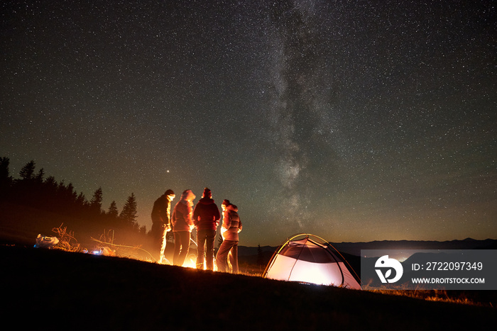 Back view group of four young friends hikers having a rest together around bonfire beside camp and i