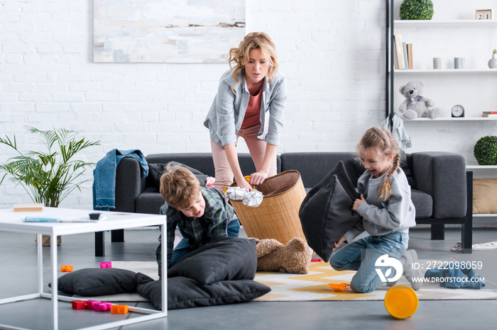 emotional tired mother putting toys in basket while naughty kids fighting with pillows