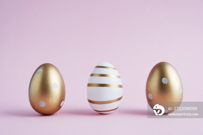 Three Easter eggs painted with gold paint in different patterns stand on a pink pastel color. Easter