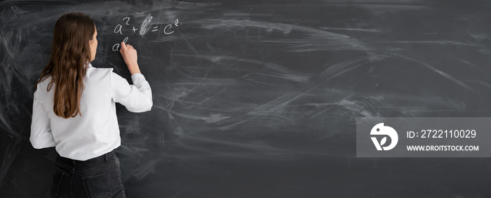 student woman or teacher in the class writes math formulas in chalk on a black chalkboard. A young girl stands with her back to the camera. Back to school. Education concept. Banner.