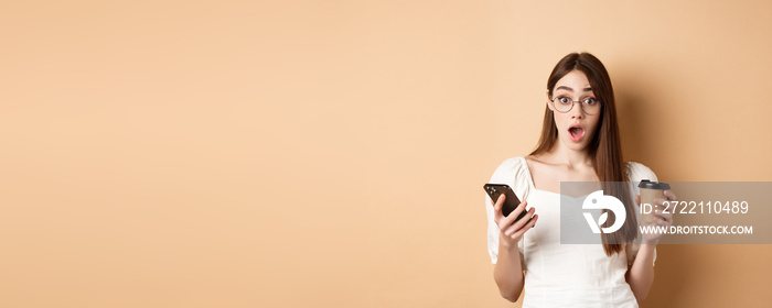 Surprised girl react to mobile phone news, holding smartphone and cup of coffee, look excited with dropped jaw at camera, beige background