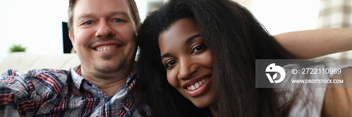 Portrait of cheerful mixed race couple taking happy selfie on sofa indoors. Man and latino woman widely smiling on camera. Multinational and biracial family concept