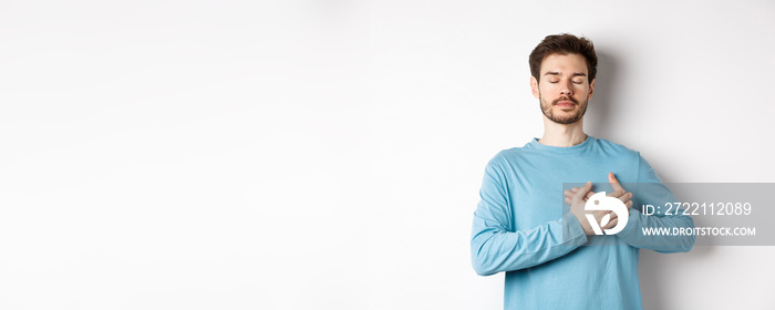 Portrait of young caucasian man with beard standing with hands on heart and closed eyes, dreaming about happy moments, standing over white background