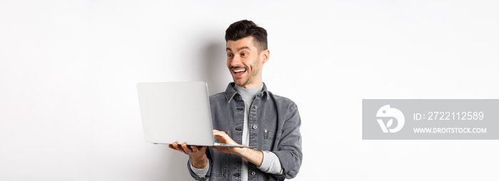 Man watching funny video on laptop, laughing at computer screen and smiling joyful, standing on white background