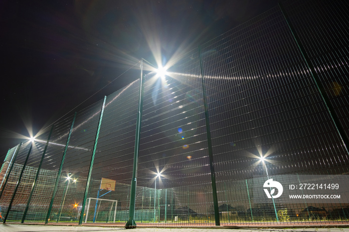 Outdoors mini football and basketball court with ball gate and basket surrounded with high protective fence brightly illuminated with spotlight lamps at night.