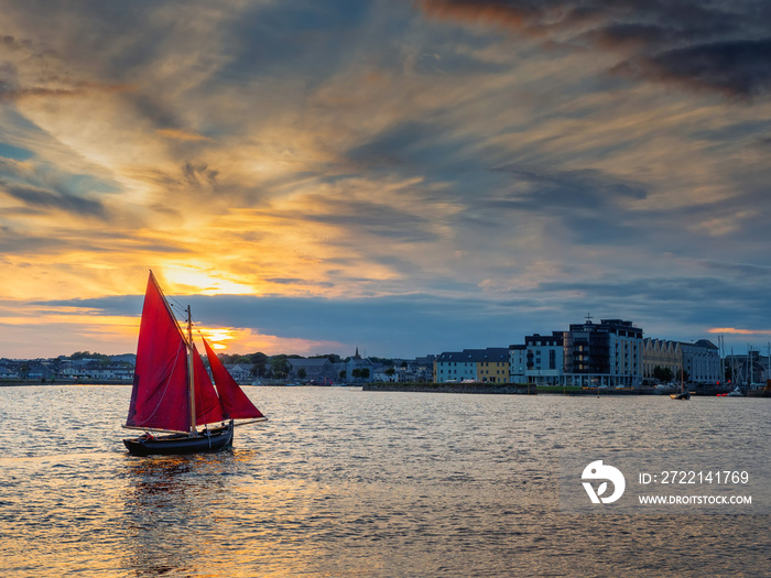 Wooden boat with red color sail going into harbor. Galway city, Ireland. Popular local type boat called Galway hooker. Sport and hobby. City building silhouette in the background. Dramatic sunset sky.