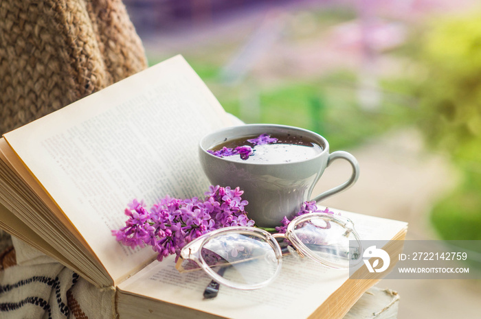 Book, glasses, cup of tea and lilac on a wooden window. Fragrant tea in the garden. Romantic concept. Vintage style