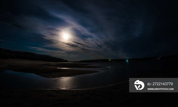 A long exposure shot of Tom river with moonlit water and shores under the cloudy night sky, foot-marks on sand.