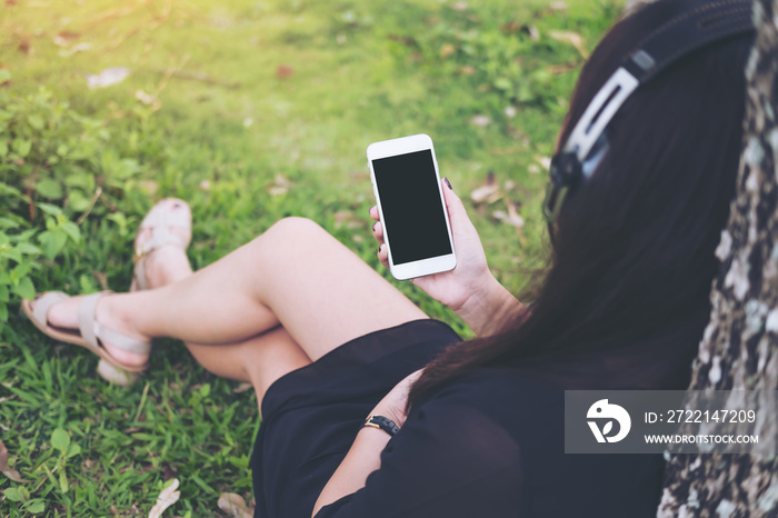 Mockup image of A beautiful Asian woman sitting and listening to music with headphone and holding smart phone by the tree with green forest background