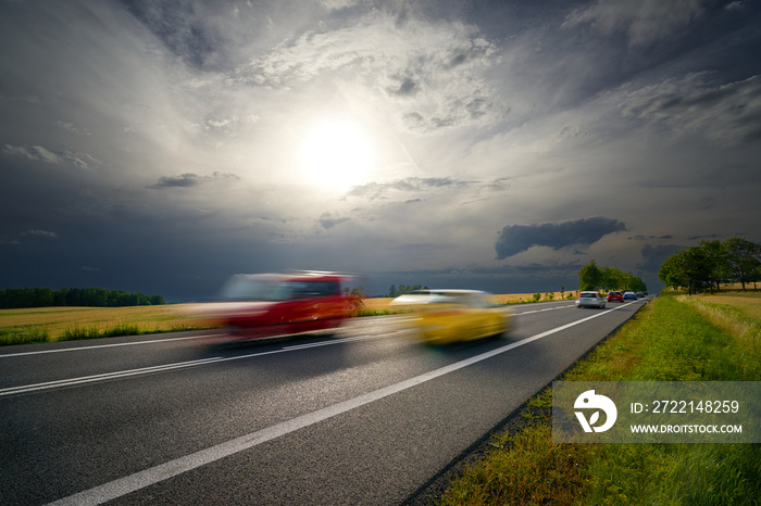 Motion blurred cars driving on a road in a rural landscape under the sun shining through the clouds before the storm