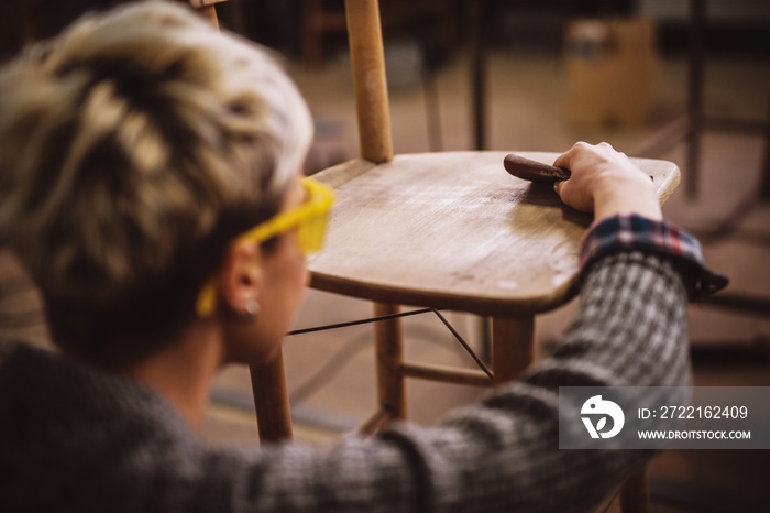 Rear view of careful professional carpenter woman sanding a chair in the workshop.