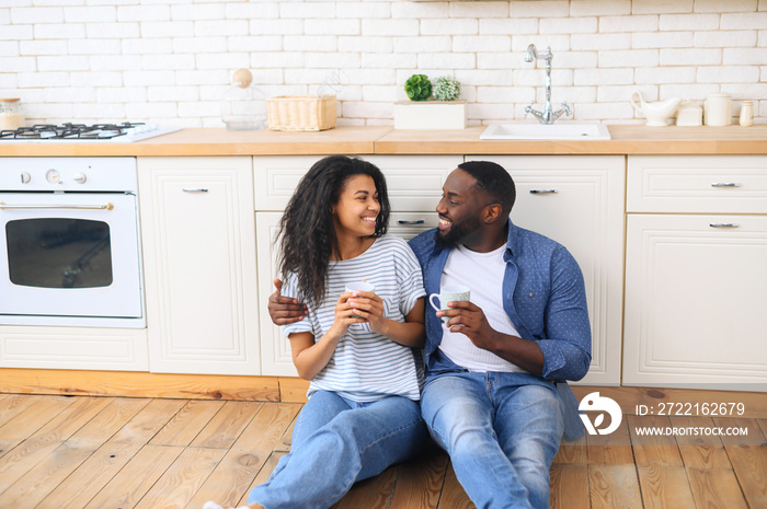 Young multiracial couple drinking coffee sitting on the kitchen floor, celebrate moving to a new apa