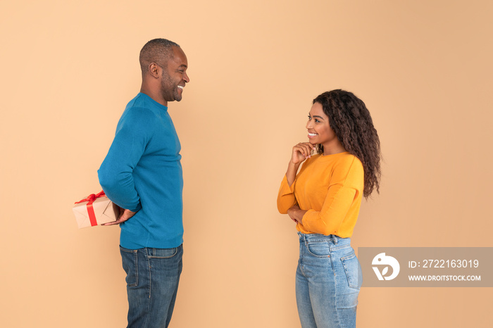 Loving african american man holding wrapped gift box, hiding present for his young wife behind back,