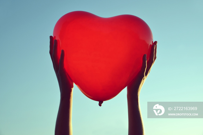 Hands holding red balloon in the shape of heart at sunset.