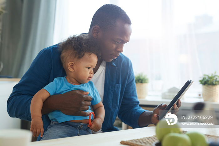 Side view portrait of African-American man holding baby while using digital tablet in home interior,