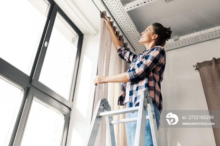 home improvement, decoration and renovation concept - happy smiling woman on ladder hanging curtains