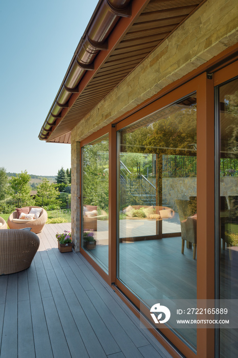 Stone garden terrace with large glass windows. Wooden floor and wicker garden furniture.