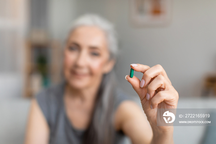 Cheerful caucasian older gray-haired lady hold pill in hand for treatment of disease in living room 