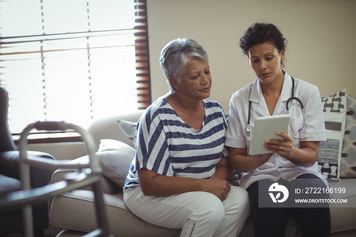 Female doctor discussing over digital tablet with senior woman in living room at home