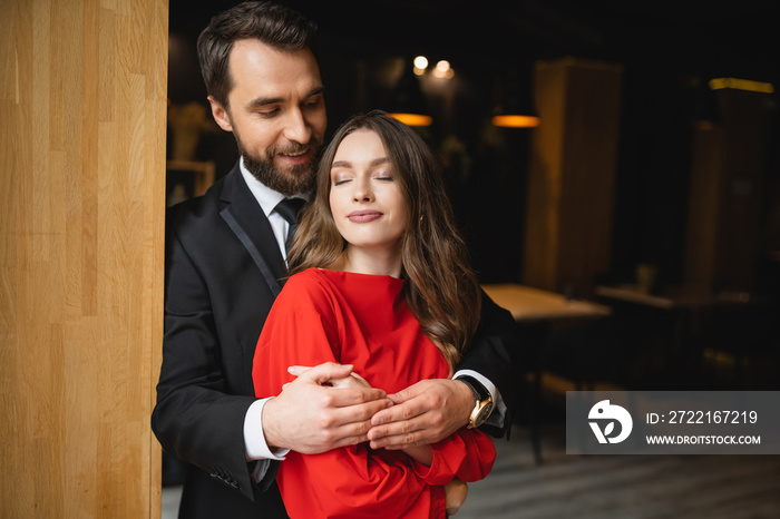 bearded man in suit hugging pleased woman in red dress on valentines day