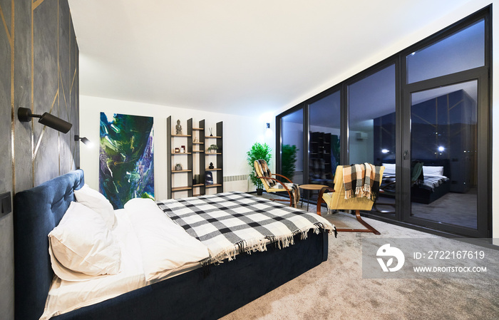 Chalet room with reflection in large panoramic window at twilight. Side view of illuminated bedroom.