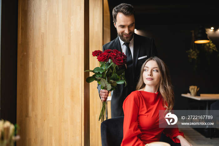 bearded man in suit smiling while holding bouquet of red roses near girlfriend on valentines day