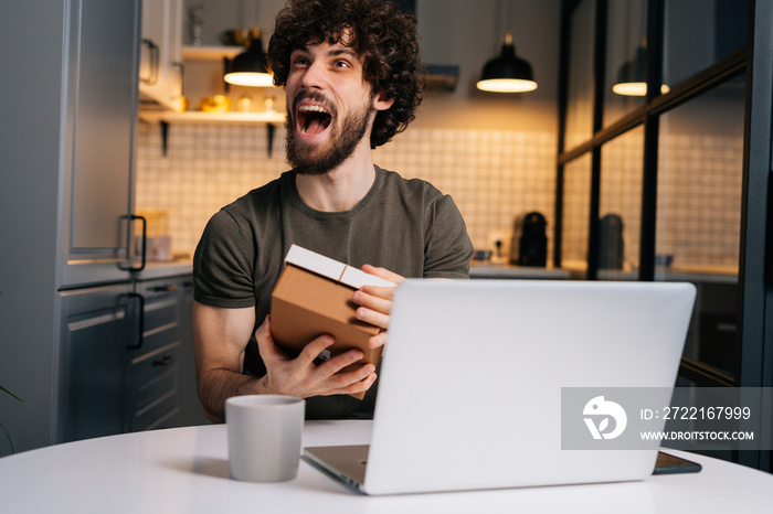 Portrait of happy excited young bearded man opening gift box with present during video call on lapto