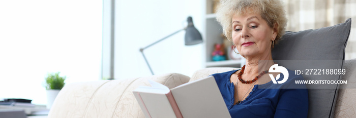 Elderly woman is reading book at home on couch